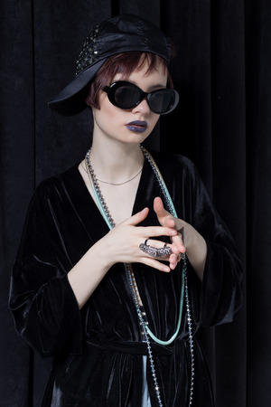 fashion shot of a beautiful young girl with a cheeky short hair in a hipster style in a black velvet jacket in Kupka Chesney and sunglasses in a studio on a black backgroundの写真素材