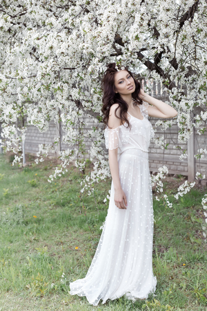 Beautiful sexy girl with a gentle make-up in a long light dress among the flowering trees walks in the spring garden on a warm sunny dayの写真素材