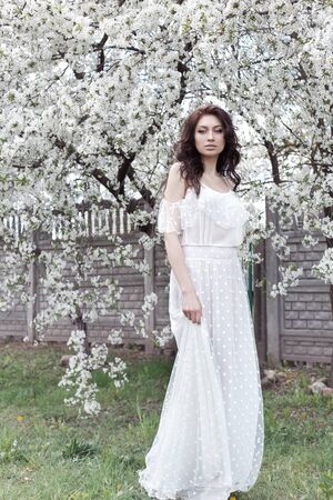 Beautiful sexy girl with a gentle make-up in a long light dress among the flowering trees walks in the spring garden on a warm sunny dayの写真素材