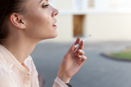 Beautiful young sad lonely girl sitting with a cigarette in hands on bench in park and smokesの写真素材