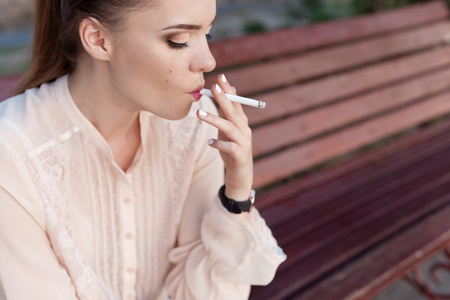 Beautiful young sad lonely girl sitting with a cigarette in hands on bench in park and smokesの写真素材