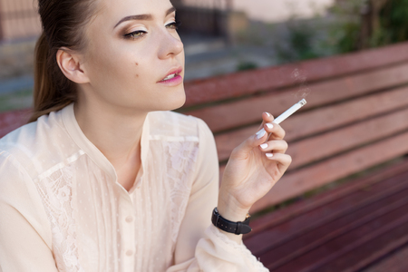 Beautiful young sad lonely girl sitting with a cigarette in hands on bench in park and smokesの写真素材