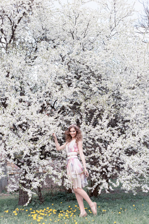 Beautiful young girl with curls in an air colored dress with a wreath on her head walking in the park near a cherry blossom on a sunny dayの写真素材