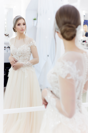 Beautiful cute tender young girl bride in wedding dress in mirrors with evening hair and gentle light make-upの写真素材