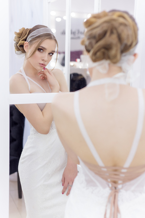 Beautiful cute tender young girl bride in wedding dress in mirrors with evening hair and gentle light make-upの写真素材