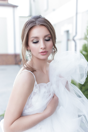 Beautiful tender sweet girl bride with a festive hairdo and light make-up with a veil in her hands with a costume jewelery earrings on a city streetの写真素材