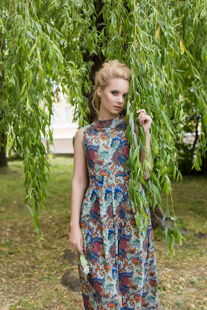Beautiful sweet tender girl blonde with blue eyes stands near a tree with long branches with a sprig of flowers in her handsの写真素材