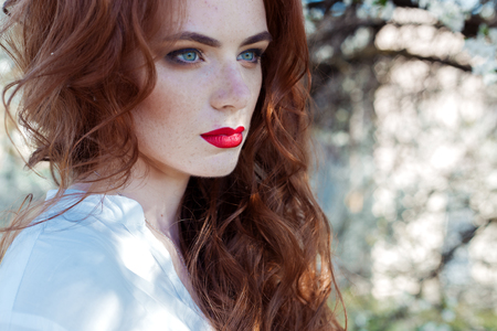 Beautiful sexy red-haired girl with freckles with red lipstick on her lips near blooming trees in the city on a sunny clear dayの写真素材