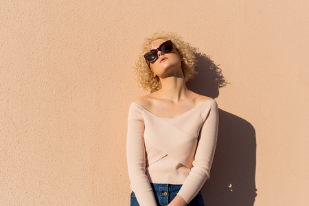 beautiful young girl with curls in sunglasses stands near the wall in sunlight at sunsetの写真素材