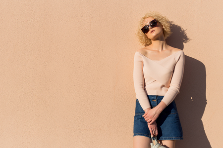 beautiful young girl with curls in sunglasses stands near the wall in sunlight at sunsetの写真素材