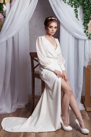 beautiful young tender girl bride in white dress with a wreath on head with blue eyes with a wedding hairstyle in a beautiful white dress with train in studioの写真素材