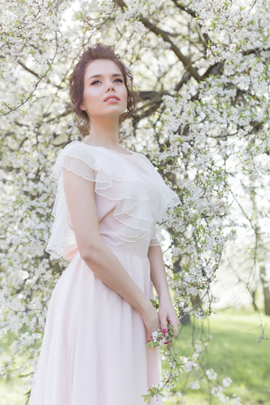 beautiful tender sweet girl in a pink dress with a hairdo near blossoming tree on a sunny spring dayの写真素材