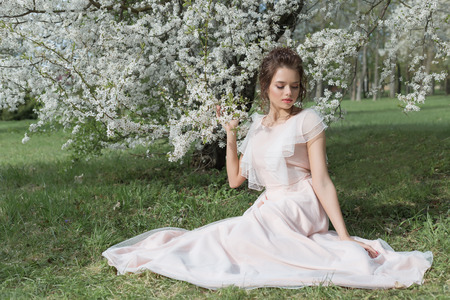 beautiful tender sweet girl in a pink dress with a hairdo near blossoming tree on a sunny spring dayの写真素材