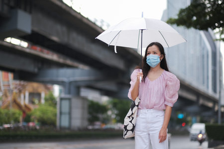 Asian woman wearing a surgical mask holds an umbrella in a rainy day to prevent the COVID-19の写真素材
