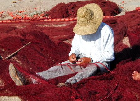 Morocco, Essaouira: fisherman reparing his fishing net; blue sky, a fisher man with his big hat seat on his red fishing net, a colorful and typical picture   の写真素材