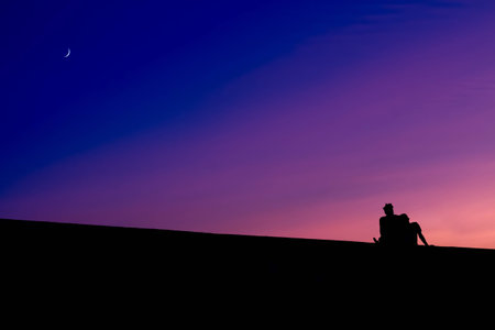 The silhouettes of two men sitting, against the purple colors of a sunset, with a slice of the moon on the blue part of the sky.の写真素材