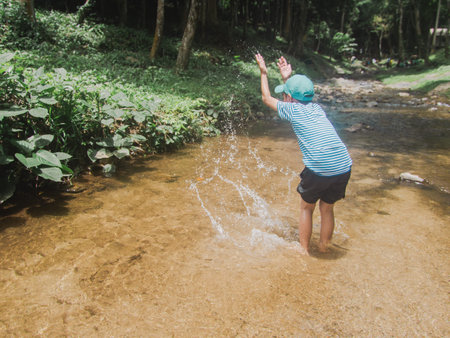A young child in a striped shirt and blue cap stands knee-deep in a clear, shallow stream, arms...