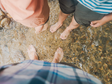 Multiple pairs of bare feet stand in a shallow, clear stream, surrounded by smooth pebbles and flowing water. The image captures a moment of simple joy and connection with nature, as friends or family members share in the refreshing sensation of cool water on a warm day, embodying the essence of outdoor exploration and sensory play.の写真素材