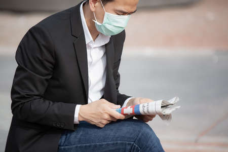 The image face of a Business Asian man wearing a doctor mask to prevent germs, toxic fumes, and COVID 19, He is reading and listening to the news on tablets and newspapers, do not work in the park.の写真素材