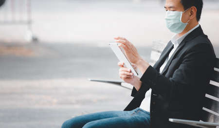 The image face of a Business Asian man wearing a doctor mask to prevent germs, toxic fumes, and COVID 19, He is reading and listening to the news on tablets and newspapers, do not work in the park.の写真素材