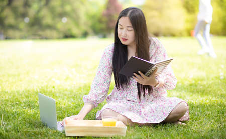Working outside in summer or spring and WFH concept, Young beautiful Asian woman reading a book and looking laptop in the natural park, Freelance working, homeschooling, distance learning.の写真素材