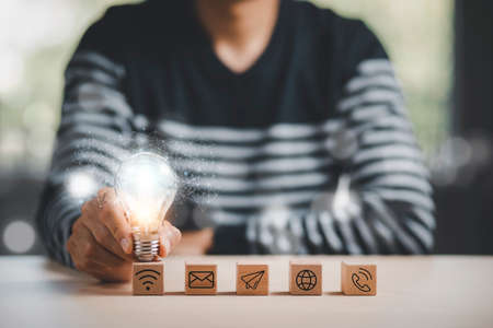 Man hand holding light bulb with Business icon on wood cube and working on desk, Creativity, and innovation are keys to success. Concept of new idea and innovation with Brain and light bulbs.の写真素材
