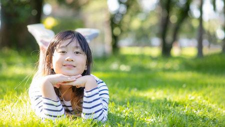 Asian young beautiful child wearing comfortable dress smiling lying on green grass on the background of a green summer park.の写真素材