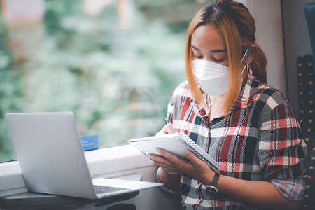 Businesswoman wearing a mask and working on laptop on a train, Asian freelance writer or designer writing emails on computer, working and smile resting on comfortable armchair near window of train.の写真素材