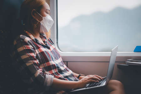 Businesswoman wearing a mask and working on laptop on a train, Asian freelance writer or designer writing emails on computer, working and smile resting on comfortable armchair near window of train.の写真素材