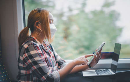 Businesswoman wearing a mask and working on laptop on a train, Asian freelance writer or designer writing emails on computer, working and smile resting on comfortable armchair near window of train.の写真素材