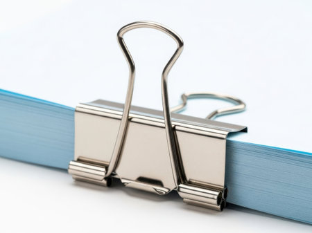 This image features a silver binder clip holding a stack of blue paper against a white background. The binder clip is prominently displayed in the center of the image, with the blue paper stack beneath it. The white background provides a clean and minimalistic backdrop, emphasizing the objects in the foreground. This image is ideal for office supply, organization, or stationery themes.の素材