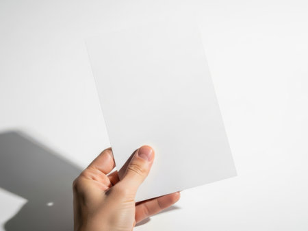 The image features a hand holding a piece of paper against a white background. The paper is slightly tilted and appears to be blank. The hand is positioned in such a way that only the fingers and thumb are visible, holding the paper from the sides. The overall scene is minimalistic and clean, emphasizing the simplicity of the paper and the hand holding it.の素材