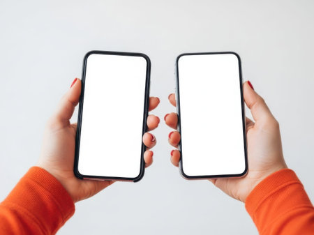 The image depicts two hands, each holding a smartphone with blank white screens. The hands are positioned in such a way that the phones are facing each other. The individuals holding the phones are wearing orange sleeves, which adds a vibrant contrast to the otherwise minimalistic scene. The background is plain and white, ensuring that the focus remains on the phones and the hands holding them. This image could symbolize technology, communication, or the digital age.の素材