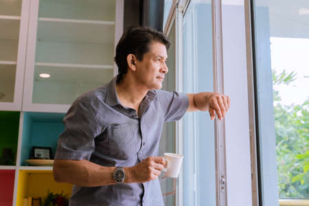 Middle-aged Asian man looking through a window, sipping coffee and using ideasの写真素材