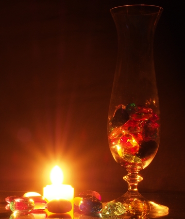 Transparent glass vase with colored pebbles. The stones in the form of hearts and flowers. Beside the candle burns. Still life.の写真素材