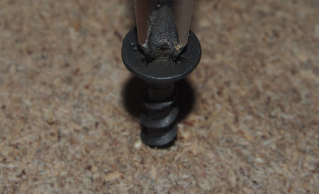 Black screw wrapped in a sheet of chipboard with a screwdriver or a drill. Close up.