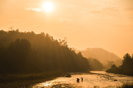 Warm toned image, silhouette of country side folks crossing river in the morning the sun ray shine through the hillsの写真素材