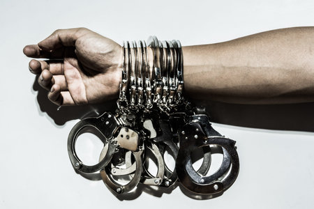 Hand wearing many handcuffs with harsh lighting set up on white wall, conceptual image of committing...