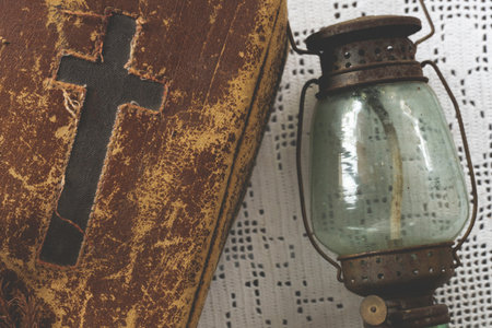 Old bible with cross on its cover on white fabric next to old rusty lampの写真素材