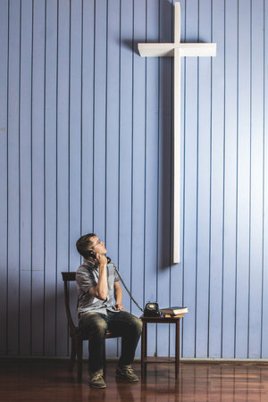 Caucasian man making a phone call looking at the white cross on wall. Concept trying to talk to godの写真素材