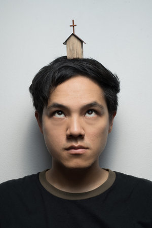Asian man looking with his eyes roll up to small wooden church on his headの写真素材