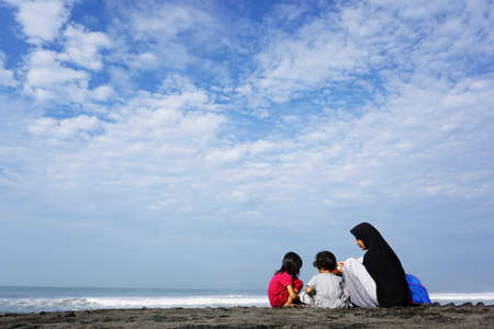 A hijabi mother plays in the sand with her two daughters on the beachの写真素材