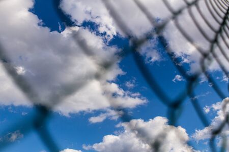 Background of Metal mesh fence against deep blue sky with white fluffy clouds backdrop. Concept of jail and freedom.の写真素材