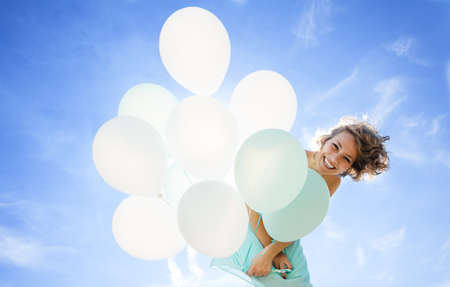 Young girl in a dress, smiling and laughing holding balloons against the skyの写真素材