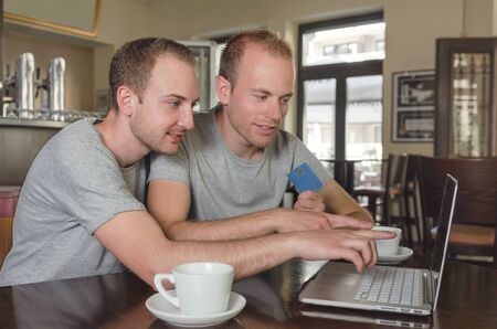 smiling gay couple holding their credit card doing online banking on their laptopの写真素材