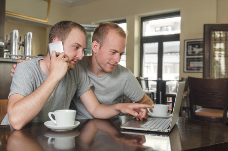 Shot of a young gays couple  speaking on a phone with laptop  in a coffee shopの写真素材