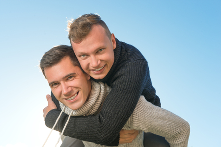 Happy young gay couple giving his  boyfriend a piggyback ride outdoors on a background of skyの写真素材