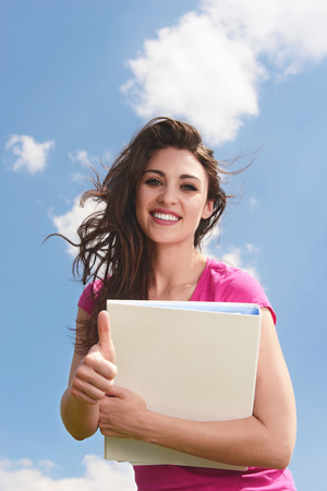 Portrait of an attractive young woman with folder smiling  brightly and giving you a thumbs upの写真素材