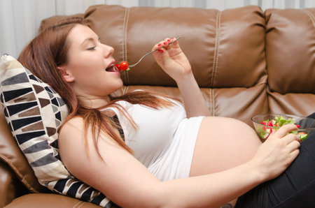 pregnant woman eating healthy salad lying in sofa