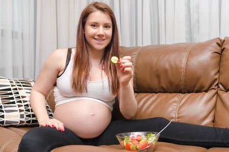 pregnant woman eating healthy salad sitting  in sofaの写真素材